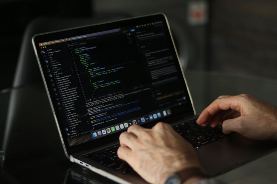 A person working on a laptop placed on a dark table, with the screen displaying lines of code in a code editor, indicating programming or digital work. The individual's hands are positioned over the k